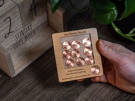 A hand holding the 10 Penny Puzzle by Creative Crafthouse, nine pennies in the wooden board's recess and the tenth set aside, next to a wooden block calendar turned to December.