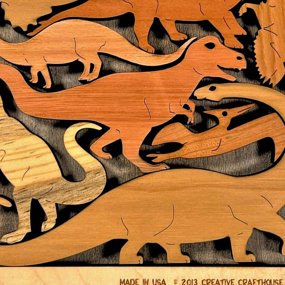 Close-up of the Dinosaur Dilemma wooden puzzle by Creative Crafthouse: dinosaur pieces in several wood tones, including a long-necked sauropod and a T. rex, above the frame's engraved edge.