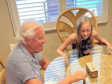 A white-haired man and a smiling girl playing Penny Drop at a round glass-top table, the wooden game box in front of them and pennies spread across the table.