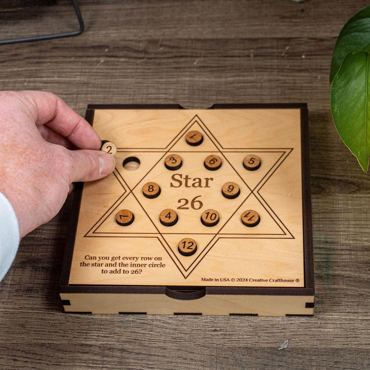 A hand lifting the number 2 disc from the Star 26 math puzzle by Creative Crafthouse, twelve numbered wooden discs on a six-pointed star board set into a wooden box on a desk.