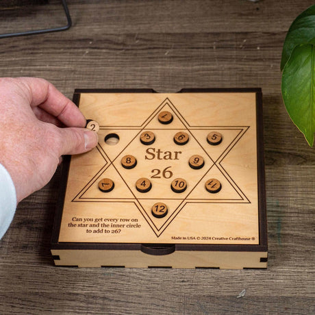 A hand lifting the number 2 disc from the Star 26 math puzzle by Creative Crafthouse, twelve numbered wooden discs on a six-pointed star board set into a wooden box on a desk.