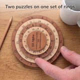 Overhead view of a hand turning the rings of the 60 Alpha puzzle on an oak table beside a pencil and a white mug. Headline: Two puzzles on one set of rings.