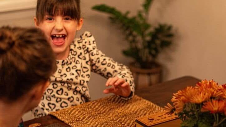 Child in a leopard print shirt excited while playing the Penny Drop game with her sister at the kitchen table.