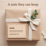 Engraved wood message card with a birthday message resting on a gift wrapped in kraft paper with a cream ribbon and eucalyptus