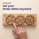 A fingertip setting a lettered gear on the Enigma II Small cipher machine, with the headline Set your three-letter keyword.