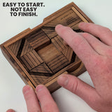 Two hands sliding the engraved raintree blocks of the Get My Goat puzzle inside its wooden tray, with the patent and maker inscription in view. Headline: Easy to start. Not easy to finish.