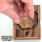 A hand lifting the engraved Get My Goat start block out of the wooden tray to open the first empty square. Headline: One block comes out to start.