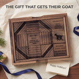 The Get My Goat wooden sliding block puzzle lying open on ivory tissue in a holiday gift scene, its engraved raintree blocks showing the octagon pattern, the goat silhouette and the USA patent 1914 maker line, with a kraft-wrapped present, indigo ribbon, evergreen sprigs and a Happy Holidays card. Headline: The gift that gets their goat.