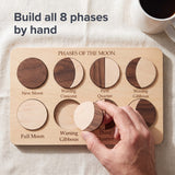 Adult hand fitting a two-tone piece into the Moon Phases Puzzle board on a linen tablecloth. Headline: Build all 8 phases by hand.