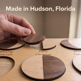 Adult hand holding a walnut half over the First Quarter recess of the Moon Phases Puzzle, its maple half already seated. Headline: Made in Hudson, Florida.