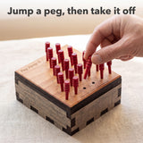 Adult hand lifting a jumped red peg off the Peg Jump 6 board after the first move. Headline: Jump a peg, then take it off.
