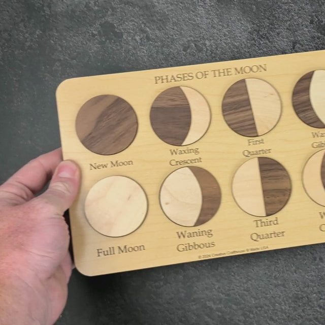 Hands build the Moon Phases Puzzle on a grey stone surface: the walnut new moon and maple full moon go in as single pieces, the other six phases from two pieces each, with the engraved learning key alongside.